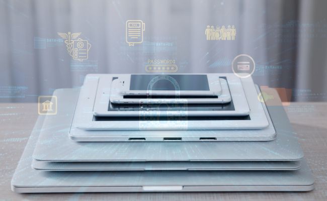 Stack of devices, laptops, tablets, cell phone with tech icons faintly in the background. 